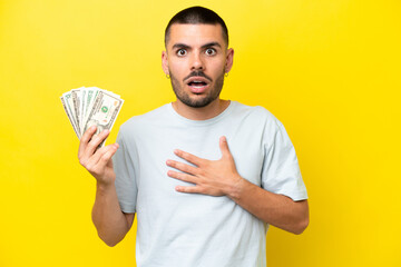 Young caucasian man taking a lot of money isolated on yellow background surprised and shocked while...