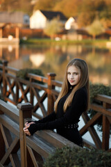 A woman stands on a wooden bridge near a picturesque pond. She leans her back against the bridge...