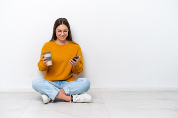 Young girl sitting on the floor isolated on white background holding coffee to take away and a...