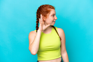 Young reddish woman isolated on blue background listening to something by putting hand on the ear