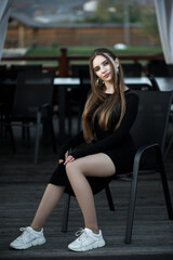 A woman sits on a black chair on a wooden terrace. She is wearing a black dress with long sleeves...