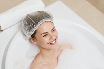 Relaxed young woman enjoying a bubble bath, wearing a shower cap, with a joyful smile on her face....