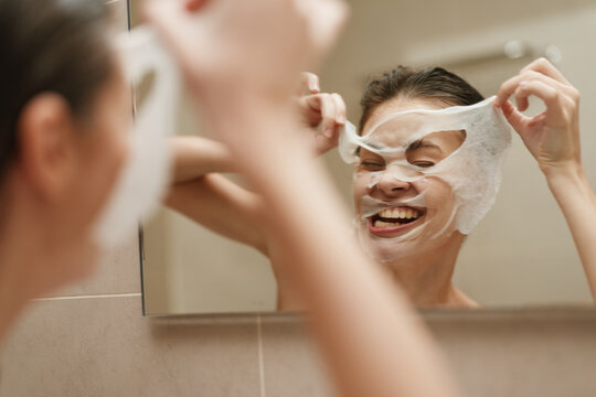 Joyful woman enjoying skincare routine at home, applying facial mask in front of mirror, promoting self-care and wellness. Healthy lifestyle concept.