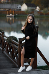 A woman stands on a wooden bridge near a picturesque pond. She leans her back against the bridge...