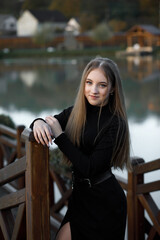 A woman stands on a wooden bridge near a picturesque pond. She leans her back against the bridge...