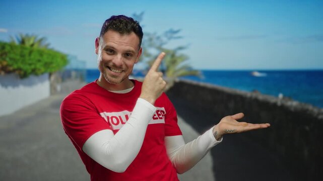 Young man in red shirt smiling and pointing with ocean view behind him along promenade with bright sky showcasing joyful and inviting seaside atmosphere for viewers