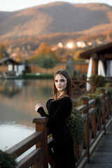 A woman leans on the railing of a wooden bridge. She has blonde hair and is wearing a black dress.
