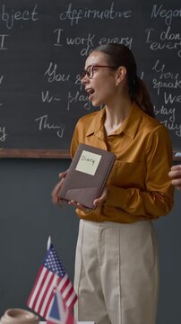 Vertical medium shot of Caucasian female instructor guiding diverse students as they use sticky notes to name and describe objects like plant, book, and headphones during lively English activity