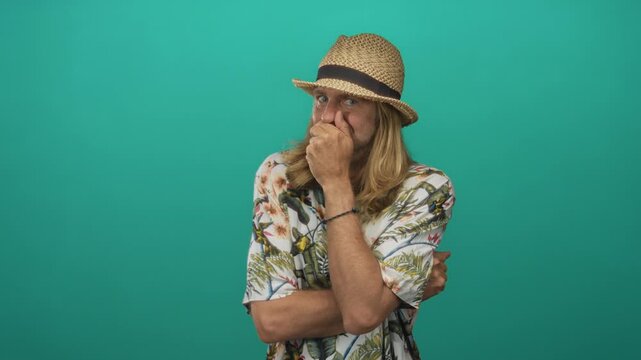 Man with long blond hair wearing straw hat and floral shirt pinches nose in teal studio with crossed arms; disgust.