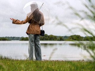Fashionable girl in brown jacket and jeans walking with see through umbrella in field by a lake....