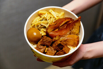 Scene of Taiwanese night market vendor preparing braised pork rice, ladling soy-braised meat and sauce into a floral paper bowl over noodles and tofu