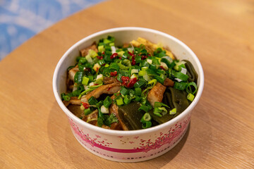 Takeout bowl of Taiwanese braised luwei featuring tofu, kelp, bean curd skin, and scallions, seasoned with chili and soy sauce, on a wooden table