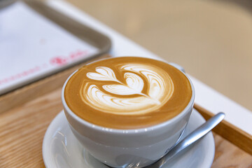 Creamy latte served in a white ceramic cup, showcasing intricate heart-shaped latte art on the velvety foam, placed on a saucer with a spoon on wooden table