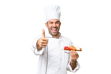 Young caucasian chef holding a sushi over isolated background with thumbs up because something good has happened