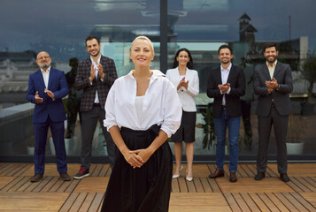 Smiling businesswoman stands on a rooftop terrace as colleagues and a diverse business team applaud...