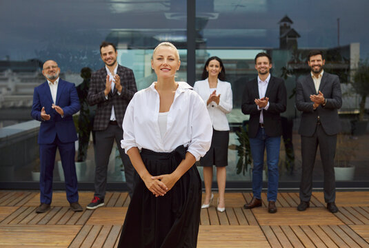 Smiling businesswoman stands on a rooftop terrace as colleagues and a diverse business team applaud her leadership in the corporate office. Business people group celebrating success. 
