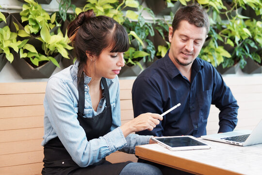 Smiling Caucasian barista couple or partnership wearing apron and consulting for marketing planning idea with tablet in the coffee shop. Start up for small cafe business owner concept. - Powered by Adobe