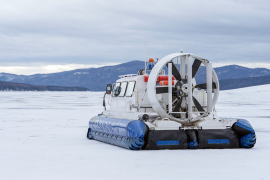 A hovercraft glides effortlessly over a snow-blanketed lake, surrounded by a serene winter landscape. The snow-covered surface reflects the cloudy sky, while distant mountains provide a picturesque
