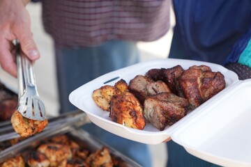 A hand uses tongs to place grilled meat into a foam takeout container at a barbecue