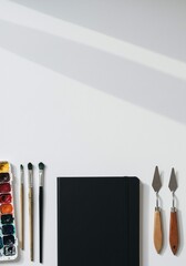 Art supplies laid out on a white surface including a palette of watercolors, paintbrushes, a black notebook, and palette knives, with soft shadows casting from the right