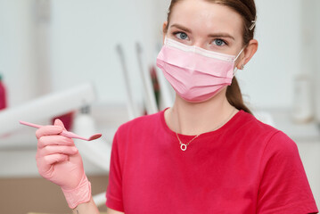 A dentist wearing a pink face mask and gloves holds a dental mirror, ready for work.