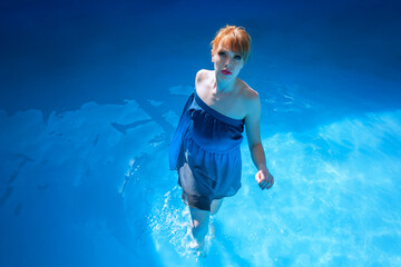Elegant Young Woman in Blue Dress Walking Through Calm Water of a Swimming Pool on a Sunny Summer...