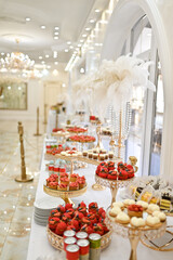 An elegant wedding reception features a rich and beautifully decorated dessert table with fruits, cakes, and sweets.