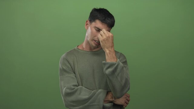 Young hispanic man pinches nose with free hand and crosses arms in green studio for stinky smell reaction; disgust.