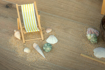 Miniature deckchair with yellow stripes surrounded by seashell and sand on wooden floor, evoking a playful beach holiday scene.