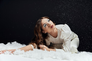 A graceful young woman in a white dress poses in falling snow with vibrant makeup.
