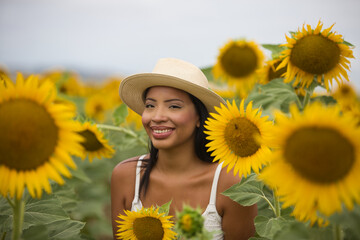 A beautiful black Latin woman wearing a white top and a straw hat stands among sunflowers. The...