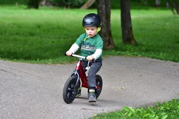 Focused toddler riding a red balance bike on a park path, wearing a green t-shirt and helmet,...