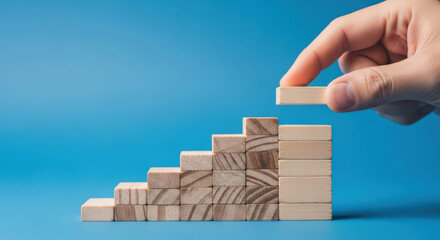 Hand Placing Wooden Block on Staircase Structure Symbolizing Step-by-Step Progress and Building Towards Success on a Blue Background for Business Growth