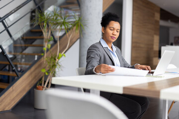 Focused businesswoman working with laptop and analyzing documents while sitting at desk in modern...