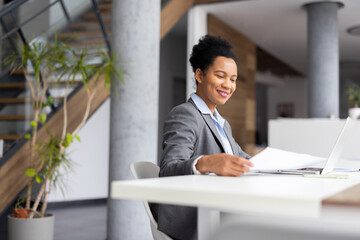 Professional businesswoman analyzing paperwork, typing on laptop, smiling in sunlit contemporary...