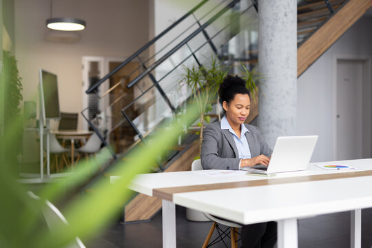 Focused businesswoman using laptop, working on project in contemporary office space with natural light and greenery