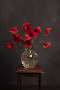 still life with red poppies in glass vase on stool in dark painterly renaissance style