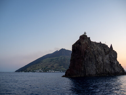 Rocks at Aeolian Island archipelago at sunset. Dark silhouettes, pink, orange and red sky and calm sea make romantic vibe.  Stromboli Island and volcano behind. 