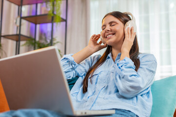 Happy relaxed Caucasian young woman in wireless headphones lying on home couch choosing listening disco music while working online on laptop netbook. Girl weekends activities entertaining. Lifestyles.