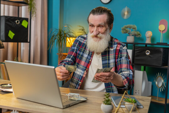 Happy Caucasian senior man counting money cash and using laptop in home office. Successful freelancer grandpa getting bundle of money in office calculates dividends earnings salary profits finances - Powered by Adobe