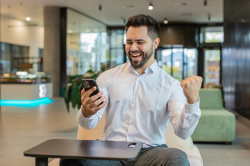 Happy young businessman guy in white shirt working on smartphone shocked by sudden victory game...