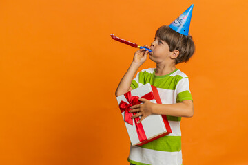 Young preteen child boy kid holding gift box wearing birthday cone hat blowing pipe celebrating...