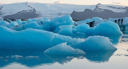 Majestic icebergs drift gracefully in a serene glacial lagoon, reflecting the muted light of an overcast sky and the distant, snow-capped mountain peaks of a pristine winter landscape