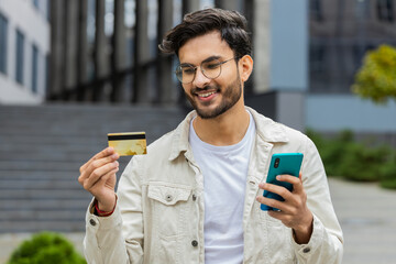 Young Indian man using credit bank card smartphone while transferring money, purchases online...