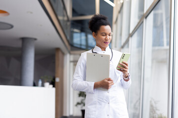 Female physician checking smartphone, carrying clipboard while walking through bright hospital hallway