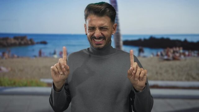 Man grins while pointing index fingers upward toward clear sky on sunny sandy beach; playful excitement.