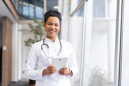 Confident female doctor smiling and holding digital tablet while standing in modern hospital corridor - Powered by Adobe