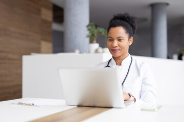Professional female physician typing on laptop during telehealth consultation, accessing patient records in contemporary medical setting