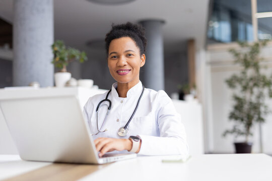 Confident female doctor using laptop, managing patient files and medical records in bright hospital office