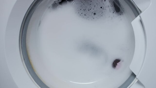 Washing clothing in domestic washing machine. Close up video of spinning washing machine drum with foam spraying door